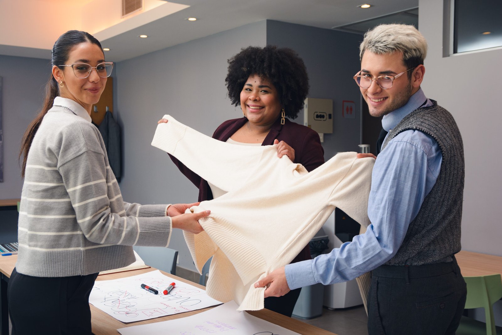 The Team of two Young Latin Women and one Man are looking at the camera smiling while working, are standing in the office happily discussing type of fabric for the clothes they are making.